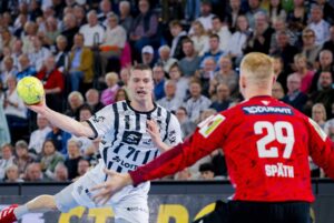 Ein Handballspieler in einem schwarz-weißen Trikot bereitet sich darauf vor, den Ball zu werfen, während ein Torwart in einem roten Trikot mit der Aufschrift 29 SPAETH ihm gegenübersteht. Die Aktion findet vor einer dicht gedrängten, jubelnden Menge statt.