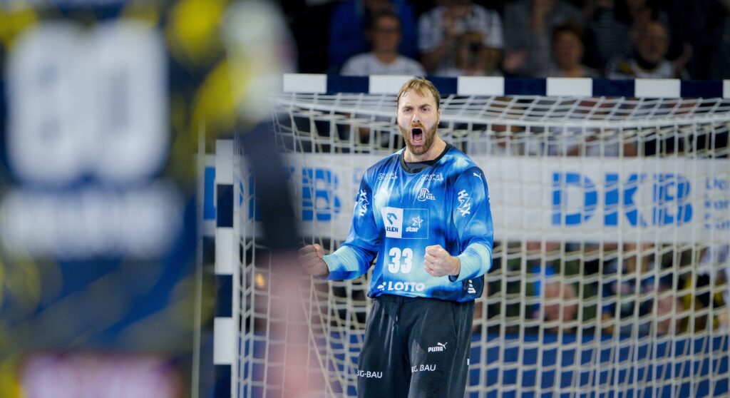 Ein männlicher Handballtorwart in einem blauen Trikot schreit und ballt die Fäuste beim Jubel vor dem Netz während eines Hallenspiels, mit verschwommenen Spielern und Zuschauern im Hintergrund.