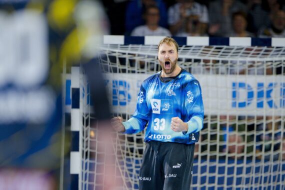 Ein männlicher Handballtorwart in einem blauen Trikot schreit und ballt die Fäuste beim Jubel vor dem Netz während eines Hallenspiels, mit verschwommenen Spielern und Zuschauern im Hintergrund.