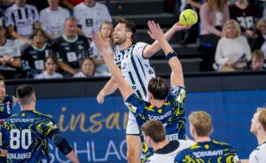 Ein Handballspieler in einem weiß-schwarzen Trikot springt, um den Ball zu werfen, während die gegnerischen Spieler in blauen und gelben Trikots die Arme heben, um ihn zu blockieren. Die Zuschauer verfolgen das Geschehen von der Tribüne aus.