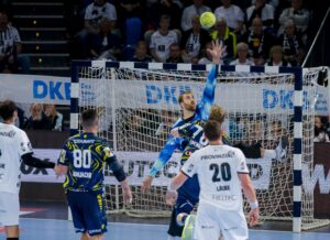 Ein Handballtorwart in Blau springt mit einem ausgestreckten Arm, um einen Schuss abzuwehren, während Spieler in weißen und blauen Trikots zusehen. Die Aktion findet vor einer überfüllten Arena und einem Tornetz statt.