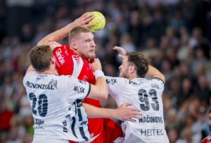 Ein männlicher Handballspieler in einem roten Trikot springt, um den Ball zu werfen, während zwei Gegner in weißen Trikots versuchen, ihn während eines Spiels in einer Halle mit einem verschwommenen Publikum im Hintergrund zu blockieren.
