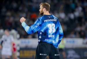 Ein Handballspieler in einem blauen Trikot mit der Nummer 33 ist von hinten zu sehen, wie er auf einem Hallenplatz jubelnd die Faust hebt, während die Zuschauer und gegnerischen Spieler im Hintergrund verschwimmen.