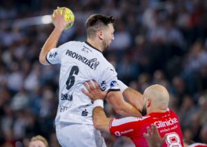 Ein Handballspieler in einem weißen Trikot springt, um den Ball zu schießen, während ein Verteidiger in einem roten Trikot versucht, ihn während eines Spiels zu blockieren, mit einer verschwommenen Menschenmenge im Hintergrund.