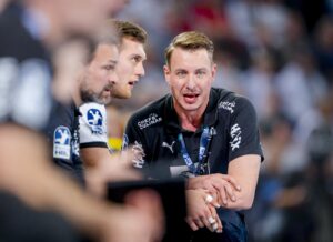 Eine Gruppe von Männern in passenden schwarzen Sportuniformen sitzt eng beieinander, konzentriert sich und spricht, möglicherweise um eine Strategie für ein Sportereignis zu entwickeln. Der vorderste Mann scheint ein Trainer oder Mannschaftsführer zu sein.