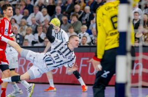 Ein Handballspieler in einem weiß-schwarz gestreiften Trikot springt und wirft den Ball in Richtung Tor, während ein gelb gekleideter Torwart sich darauf vorbereitet, den Ball abzublocken, während die Zuschauer im Hintergrund aufmerksam zusehen.