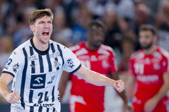 Ein männlicher Handballspieler in einem schwarz-weiß gestreiften Trikot jubelt leidenschaftlich auf dem Spielfeld und ballt die Fäuste, während im Hintergrund zwei Gegner in roten Trikots stehen.