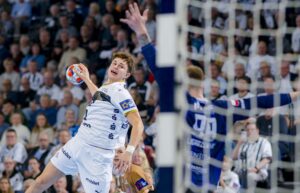 Ein Handballspieler in einem weißen Trikot springt zum Wurf, während ein Verteidiger in blauer Uniform versucht, den Ball zu blocken. Das Spiel findet in einer überfüllten Halle statt, in der die Zuschauer aufmerksam zusehen.