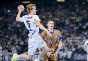 Ein männlicher Handballspieler in einem weißen Trikot springt, um den Ball zu werfen, während ein Spieler in einem braunen Trikot vor ihm steht. Sie befinden sich auf einem Hallenplatz, während im Hintergrund eine Menschenmenge zuschaut.