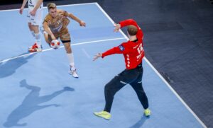 Ein Handballspieler in einem goldenen Trikot springt, um den Ball in Richtung Tor zu schießen, während ein Torwart in einem roten Trikot und einer schwarzen Hose seine Arme und Beine ausbreitet, um den Schuss auf einem blauen Spielfeld zu blockieren.