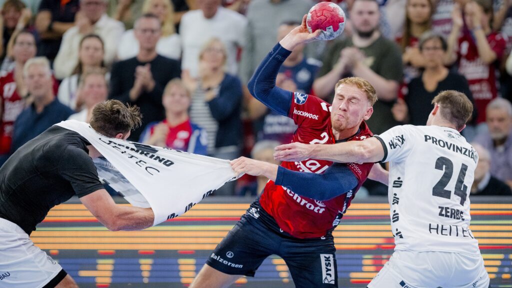 Ein Handballspieler in einem rot-blauen Trikot versucht zu schießen, während er von zwei weiß gekleideten Spielern, von denen einer an seinem Trikot zerrt, aggressiv verteidigt wird. Eine Menschenmenge beobachtet das intensive Geschehen im Hintergrund.