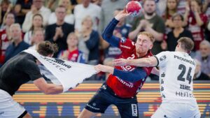 Ein Handballspieler in einem rot-blauen Trikot versucht zu schießen, während er von zwei weiß gekleideten Spielern, von denen einer an seinem Trikot zerrt, aggressiv verteidigt wird. Eine Menschenmenge beobachtet das intensive Geschehen im Hintergrund.