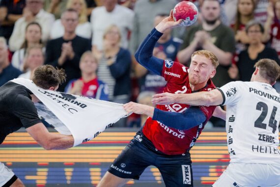 Ein Handballspieler in einem rot-blauen Trikot versucht zu schießen, während er von zwei weiß gekleideten Spielern, von denen einer an seinem Trikot zerrt, aggressiv verteidigt wird. Eine Menschenmenge beobachtet das intensive Geschehen im Hintergrund.