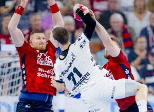 Drei männliche Handballspieler sind während eines Spiels in Aktion. Der Spieler in Weiß versucht zu schießen, während zwei Spieler in Rot eng anliegend verteidigen und die Arme heben. Die Szene ist intensiv, und die Zuschauer sind im Hintergrund verschwommen.