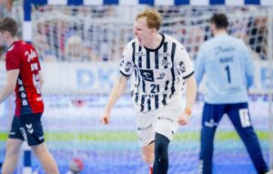 Ein Handballspieler in einem schwarz-weiß gestreiften Trikot jubelt ausgelassen auf dem Spielfeld. Im Hintergrund sind andere Spieler und ein Tor zu sehen. Ein Spieler in Rot geht weg und ein Torwart in Hellblau steht neben dem Tor.