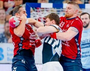 Zwei männliche Handballspieler in roten Trikots blockieren aggressiv einen männlichen Gegenspieler in einem weiß-schwarzen Trikot und packen ihn an Armen und Hals, als er versucht, den Ball zu spielen. Die Szene wirkt intensiv und körperlich.