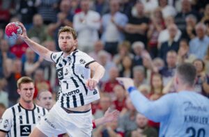 Ein Handballspieler in einem weiß-schwarzen Trikot springt, um den Ball zu werfen, und zielt auf das Tor, während der Torwart in Blau sich darauf vorbereitet, den Ball abzuwehren. Die Zuschauer auf der Tribüne im Hintergrund schauen aufmerksam zu.