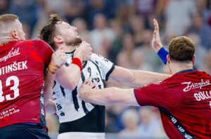 Drei männliche Handballspieler in Aktion; zwei Spieler in roten Trikots verteidigen gegen einen Spieler in einem weiß-schwarzen Trikot, der sich zu wehren scheint, während er gepackt und geblockt wird. Der Hintergrund ist mit Zuschauern unscharf.
