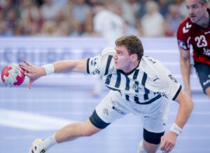Ein männlicher Handballspieler in einem weiß-schwarzen Trikot stürzt nach vorne und greift nach einem roten Ball. Ein anderer Spieler in einem roten Trikot ist im Hintergrund auf einem blauen Hallenplatz zu sehen.