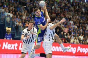 Ein Handballspieler in einem blau-grünen Trikot springt, um den Ball zu schießen, während er während eines Spiels von Spielern in weißen und schwarzen Trikots eng verteidigt wird.