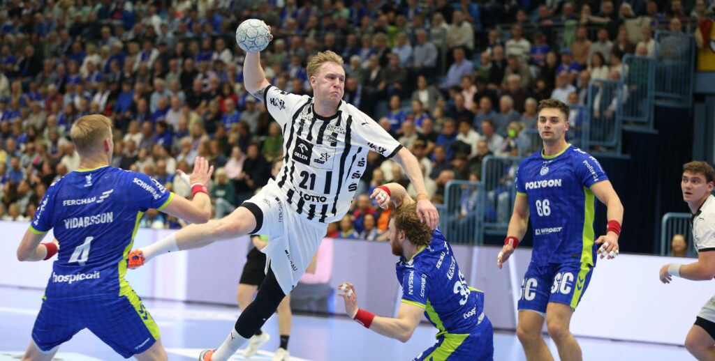 1. DAIKIN HBL  Bundesliga, VfL Gummersbach - THW Kiel Ein Handballspieler in einem weißen Trikot springt zum Wurf, während blau gekleidete Verteidiger versuchen, ihn zu blockieren. Das Spielfeld ist von einem dicht gedrängten Publikum und hellen Lichtern umgeben.