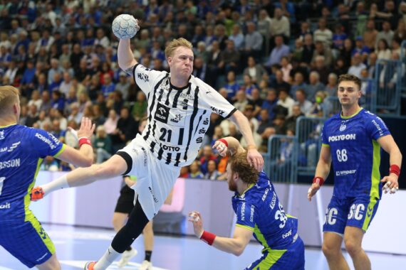 Ein Handballspieler in einem weißen Trikot springt zum Wurf, während blau gekleidete Verteidiger versuchen, ihn zu blockieren. Das Spielfeld ist von einem dicht gedrängten Publikum und hellen Lichtern umgeben.