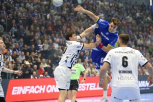 Ein Handballspieler in einem blauen Trikot springt, um den Ball zu schießen, während zwei Spieler in weißen Trikots versuchen, ihn während eines Spiels in einer überfüllten Halle zu blockieren.