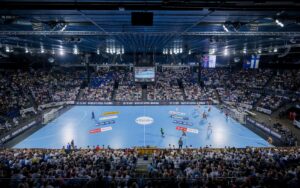 Eine große, mit Zuschauern gefüllte Halle, in der ein Handballspiel stattfindet. Zwei Mannschaften spielen auf einem blauen Spielfeld, darüber sind Anzeigetafeln und Fahnen zu sehen. Helle Lichter beleuchten die Spieler und die jubelnde Menge.