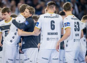 Eine Gruppe männlicher Handballspieler in weißen Trikots steht zusammen; zwei von ihnen umarmen sich, während andere mit dem Gesicht zur Seite ihre Namen und Nummern zeigen, darunter Reinkind (6) und Bilyk (53). Die Atmosphäre scheint emotional zu sein.