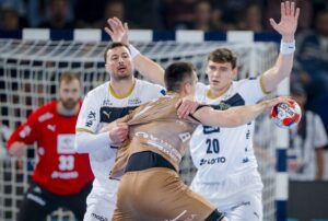 Ein Handballspieler in einem braunen Trikot versucht, zwei Verteidiger in weißen Trikots zu überwinden, während ein Torwart in Rot vom Tor aus im Hintergrund zusieht. Die Szene ist angespannt und actionreich.