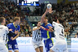 Eine Gruppe männlicher Handballspieler aus zwei Mannschaften, eine in Blau und eine in Weiß, kämpft in einer überfüllten Halle mit einem großen Publikum im Hintergrund intensiv um den Ball.