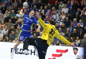 Ein Handballspieler in Blau springt, um den Ball zu werfen, während ein Torwart in Gelb versucht, den Schuss zu blocken. Beide sind in Aktion, umgeben von Zuschauern in einer überfüllten Halle.