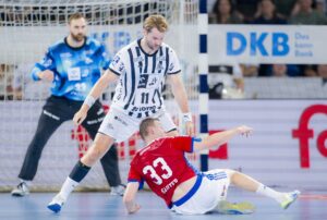 Ein Handballspieler in einem weiß-schwarz gestreiften Trikot steht über einem Gegner in einem roten Trikot, der auf das Spielfeld gefallen ist, während im Hintergrund ein Torwart und ein Tor zu sehen sind.