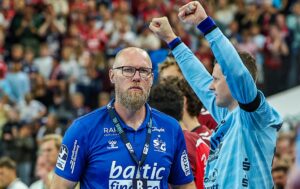 Ein Trainer in einem blauen Hemd mit Brille und Schlüsselband steht mit ernster Miene auf einem Sportplatz, während ein Spieler in einem hellblauen Trikot im Hintergrund beide Fäuste hebt. Eine Menschenmenge und andere Spieler sind ebenfalls zu sehen.