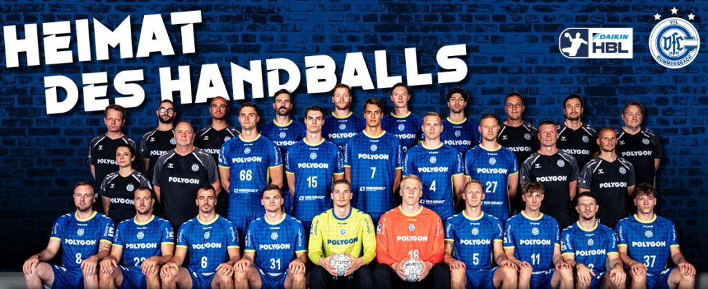 25_vor_Gumm_Teambild Eine Handballmannschaft stellt sich in drei Reihen vor einer dunklen Backsteinwand auf. Die meisten Spieler tragen blaue Trikots, einige wenige sind schwarz und zwei orange. Oben auf den Trikots stehen