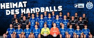 Eine Handballmannschaft stellt sich in drei Reihen vor einer dunklen Backsteinwand auf. Die meisten Spieler tragen blaue Trikots, einige wenige sind schwarz und zwei orange. Oben auf den Trikots stehen "Heimat des Handballs" und Logos.