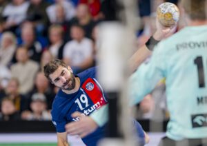 Ein Handballspieler in einem blau-roten Trikot mit der Nummer 19 befindet sich mitten im Wurf und zielt auf das Tor, während der Torwart in einem hellgrünen Trikot versucht, den Schuss zu parieren. Die Zuschauer sind im Hintergrund unscharf zu erkennen.