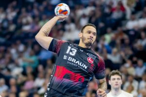Ein männlicher Handballspieler, der ein schwarz-rotes Trikot mit der Nummer 13 trägt, bereitet sich während eines Spiels auf den Wurf vor, während im Hintergrund eine unscharfe Menschenmenge zu sehen ist.