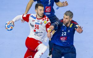 Zwei männliche Handballspieler sind auf einem Hallenplatz in Aktion; der eine in einem rot-weißen Trikot mit dem Ball, der andere in einem blauen Trikot bei der Verteidigung. Beide wirken konzentriert und intensiv.