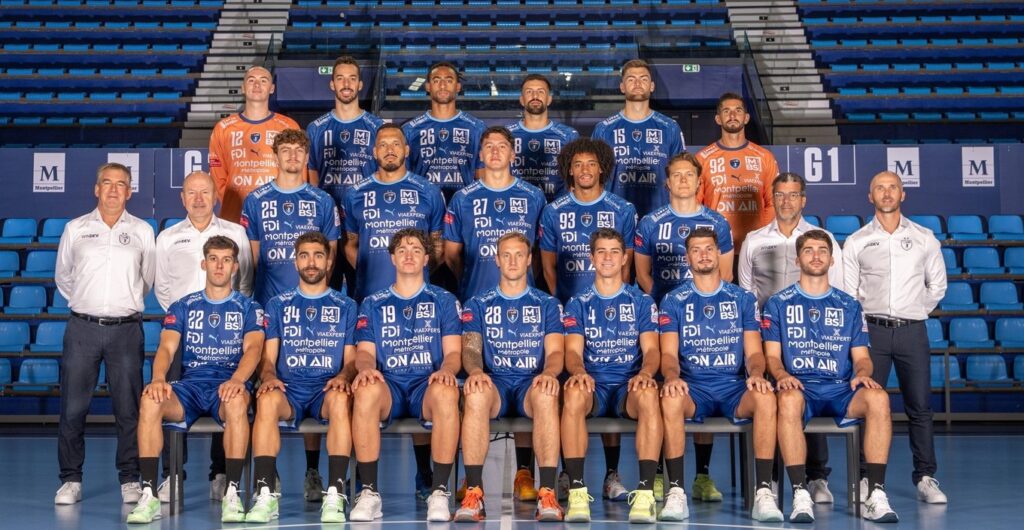 25_vor_MHB_Teambild Eine Herren-Handballmannschaft stellt sich für ein Gruppenfoto in einem Stadion auf. Die Spieler in blauen Trikots sitzen und stehen, flankiert von Trainern in weißen Hemden und leeren blauen Sitzen im Hintergrund.