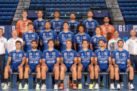 Eine Herren-Handballmannschaft stellt sich für ein Gruppenfoto in einem Stadion auf. Die Spieler in blauen Trikots sitzen und stehen, flankiert von Trainern in weißen Hemden und leeren blauen Sitzen im Hintergrund.