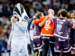 Ein niedergeschlagener Handballspieler in Weiß bedeckt seinen Kopf mit seinem Trikot, während eine Gruppe von Spielern in Lila im Hintergrund auf einem Spielfeld feiert.