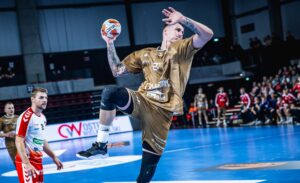 Ein Handballspieler in einem goldenen Trikot springt in die Luft, um den Ball zu werfen, während andere Spieler in roten und weißen Trikots während eines Spiels in einer Halle zusehen.