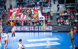 Ein Handballspiel in einer überfüllten Halle; die Fans halten ein großes Banner mit einem Löwen in einem rot-weißen Trikot. Die Spieler in rot-weißen und goldenen Trikots stehen auf dem blauen Spielfeld. Auf einem Schild steht "GROSSARTIGKEIT WIRD VERDIENKT".