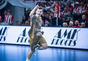 Ein männlicher Handballspieler in einem braunen Trikot jubelt mit erhobener Faust, während er über das Spielfeld rennt, während im Hintergrund begeisterte Zuschauer und Mannschaftsfahnen zu sehen sind.