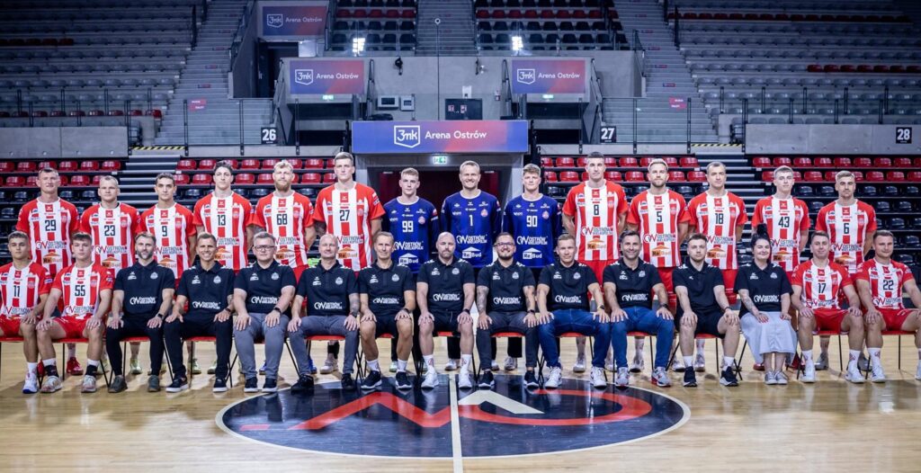 25_vor_Ostrow_Team Eine Herrenhandballmannschaft posiert für ein Gruppenfoto in einer Halle. Die Spieler tragen rot-weiße oder blaue Trikots, während das Personal in schwarzen Hemden vorne sitzt. Im Hintergrund sind leere Stadionsitze zu sehen.