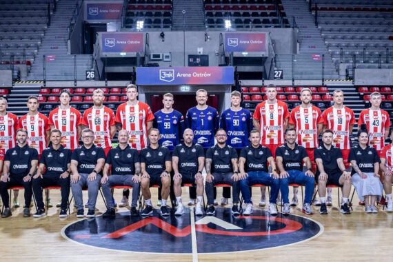 Eine Herrenhandballmannschaft posiert für ein Gruppenfoto in einer Halle. Die Spieler tragen rot-weiße oder blaue Trikots, während das Personal in schwarzen Hemden vorne sitzt. Im Hintergrund sind leere Stadionsitze zu sehen.