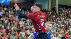 Ein Handballspieler in einem roten Trikot springt zum Wurf, während ein Verteidiger in Blau versucht, ihn zu blockieren, während im Hintergrund eine Zuschauermenge zusieht.