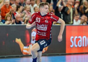 Ein männlicher Handballspieler in einem rot-schwarzen Trikot mit der Nummer 63 läuft auf einem Hallenplatz, während im Hintergrund eine jubelnde Menge und Sponsorenbanner zu sehen sind.