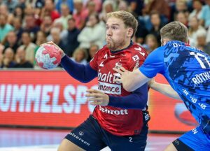 Ein männlicher Handballspieler in einem roten Trikot hält einen Ball, während er von einem Gegner in einem blauen Trikot während eines Spiels verteidigt wird, mit einer Menge von Zuschauern im Hintergrund.